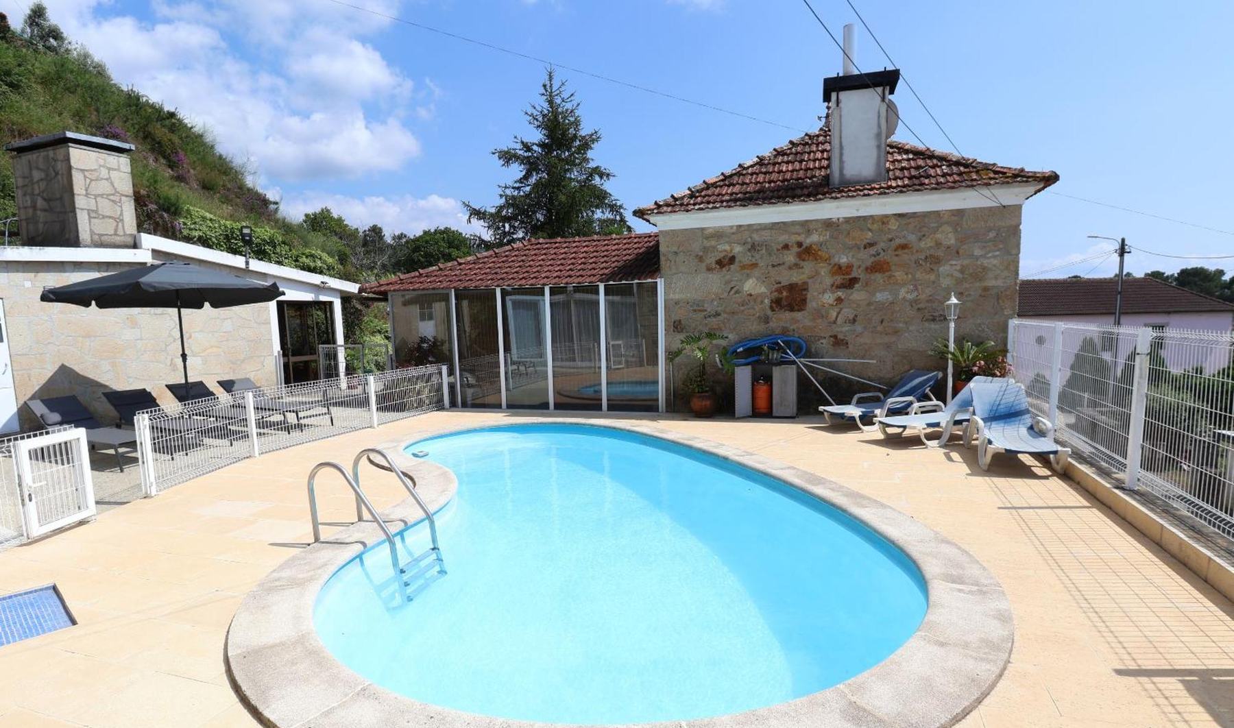 A piscina privada da Casa Pedrosa ao sol, com a casa de pedra e o telhado de telha ao fundo